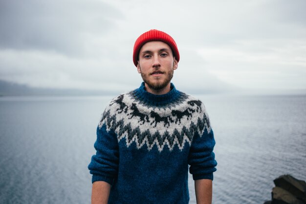 Młody atrakcyjny, brodaty mężczyzna tysiąclecia w czerwonej czapce rybaka lub marynarza i tradycyjnym islandzkim kolorze niebieskim swetrze