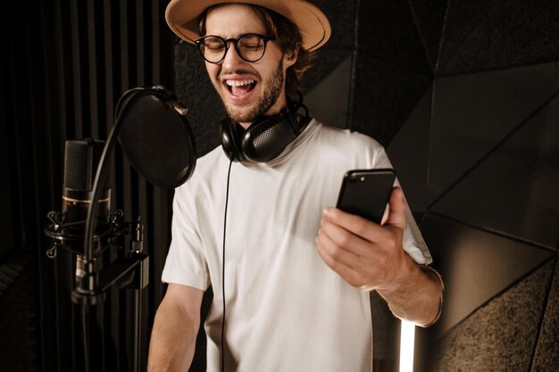 Młody atrakcyjny męski piosenkarz ze smartfonem śpiewa emocjonalnie w nowoczesnym studiu nagraniowym