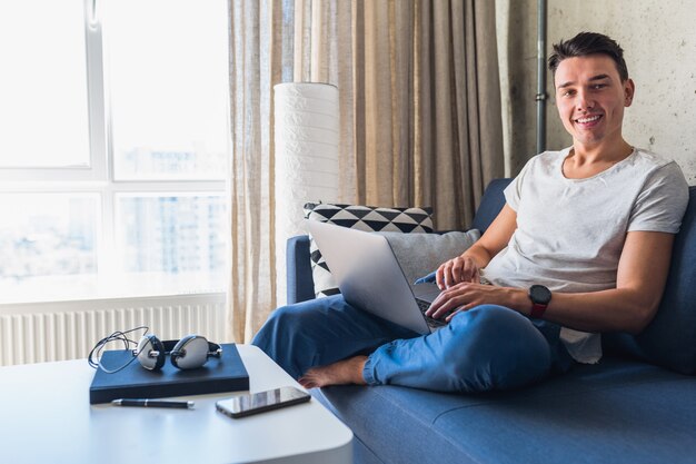 Młody atrakcyjny mężczyzna siedzi na kanapie w domu, pracując na komputerze przenośnym w trybie online, za pomocą Internetu