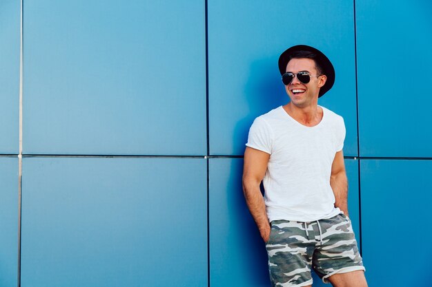 Młody atrakcyjny mężczyzna stoi na błękitnym ściennym tle w okularach przeciwsłonecznych