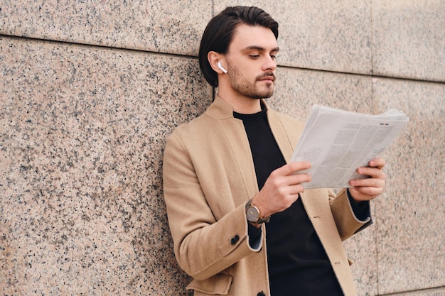 Bezpłatne zdjęcie młody atrakcyjny stylowy mężczyzna w beżowym płaszczu pewnie czyta gazetę na świeżym powietrzu