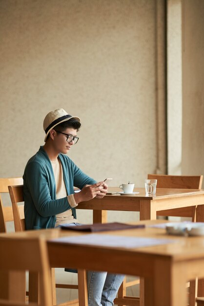 Młody Azjatycki męski modnisia obsiadanie przy stołem w kawiarni i używać smartphone