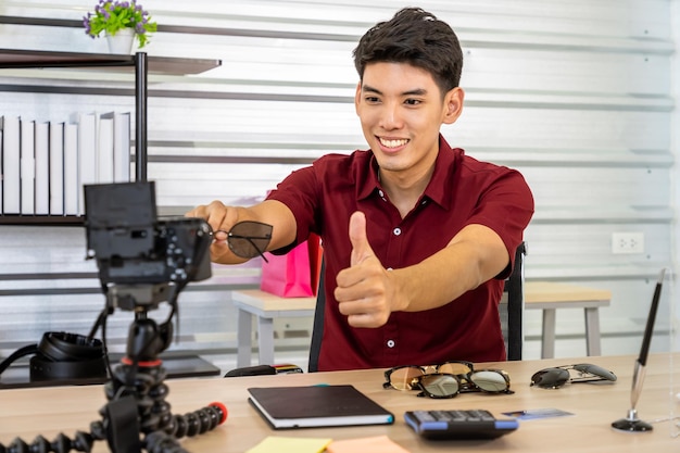 Młody azjatycki mężczyzna, bloger handlowy, który używa kamery do oglądania wideo na żywo, aby przeglądać i sprzedawać okulary. Media społecznościowe Influencer i koncepcja zakupów online