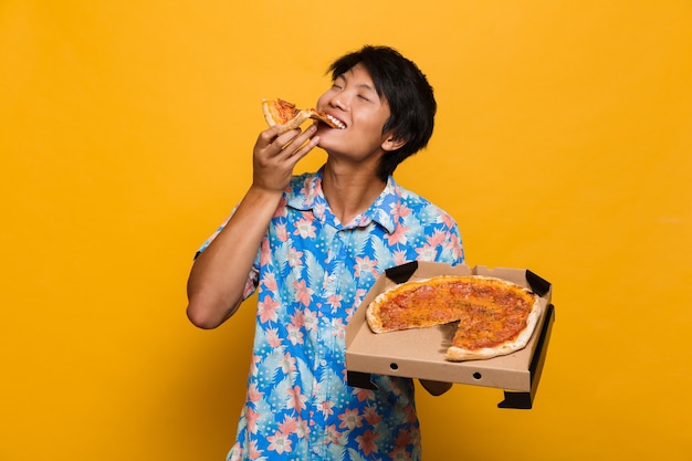 Młody azjatycki mężczyzna stojący na białym tle nad żółtą przestrzenią jeść pizzę.