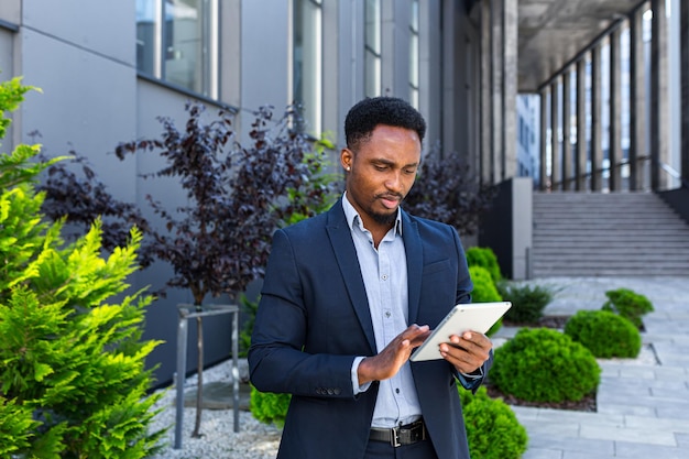 Młody biznesmen african american w formalnym garniturze stojący pracy z tabletem w ręce na tle nowoczesny biurowiec na zewnątrz. mężczyzna korzystający ze smartfona lub telefonu komórkowego na zewnątrz ulicy miasta