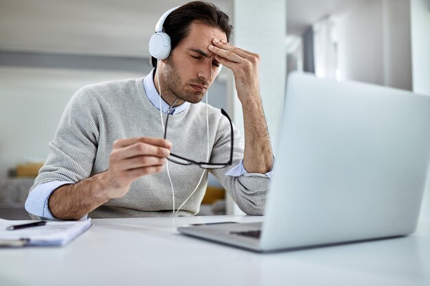Młody biznesmen ma ból głowy i czuje się wyczerpany podczas pracy na laptopie w domu