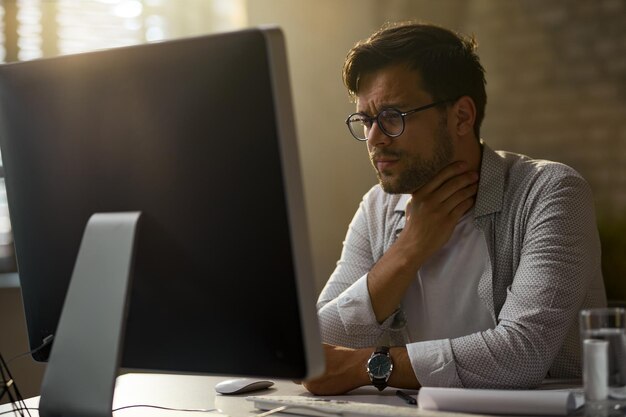 Młody biznesmen odczuwa ból w gardle podczas pracy na komputerze stacjonarnym w biurze