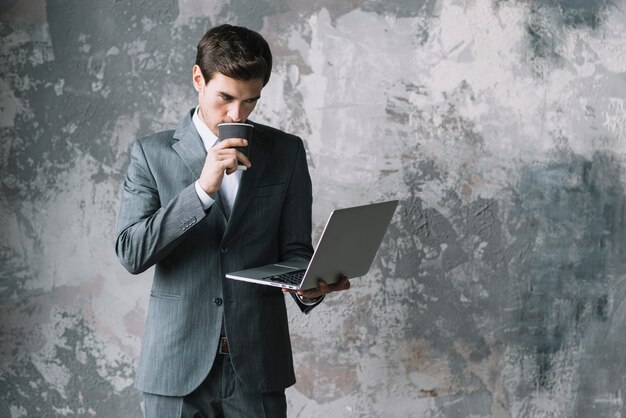 Młody biznesmen pije kawę podczas gdy patrzejący laptop przeciw starej ścianie