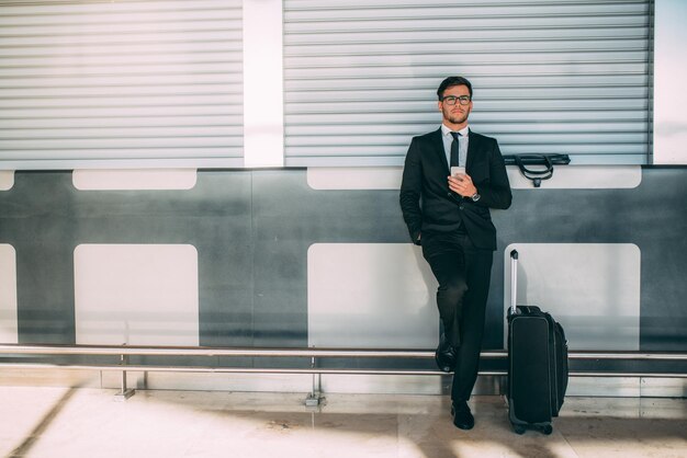Młody biznesmen stojący na telefonie z walizką na lotnisku czeka na lot xAxA