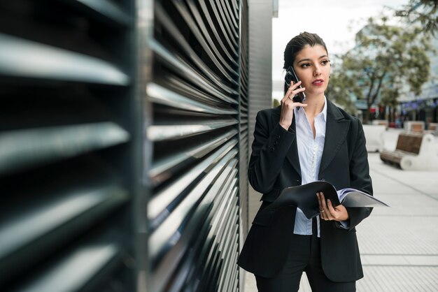 Młody bizneswoman opowiada na telefonu komórkowego mienia falcówce w ręce patrzeje daleko od