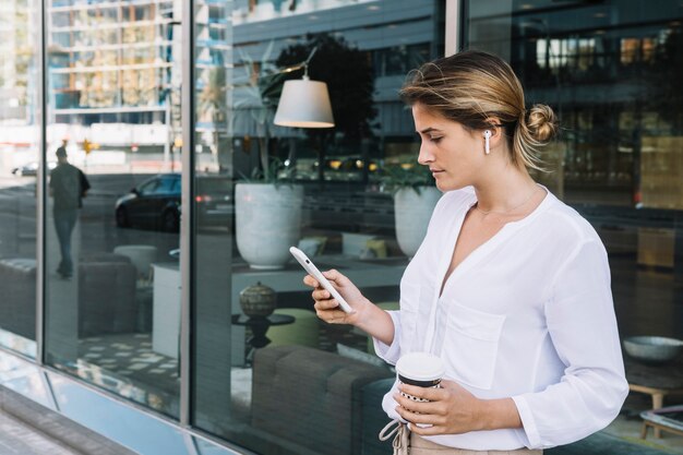 Młody bizneswoman trzyma rozporządzalną filiżankę używać telefon komórkowego