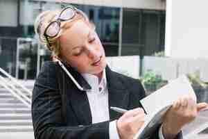 Bezpłatne zdjęcie młody bizneswomanu słuchanie na telefonu komórkowego writing na dzienniczku z piórem