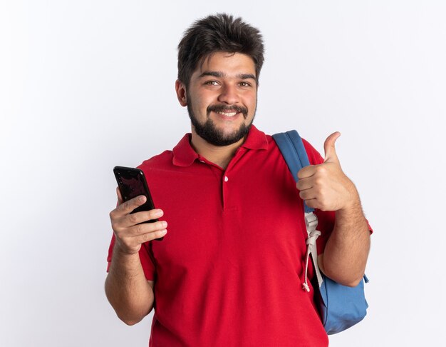 Młody, brodaty facet student w czerwonej koszulce polo z plecakiem, trzymający smartfon, uśmiechający się radośnie pokazując kciuk do góry stojący nad białą ścianą