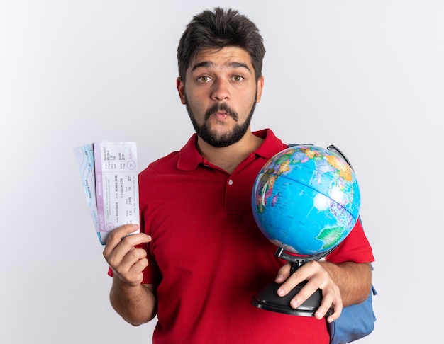 Młody, brodaty facet student w czerwonej koszulce polo z plecakiem trzymającym bilety lotnicze i kulą ziemską zaskoczony stojąc nad białą ścianą