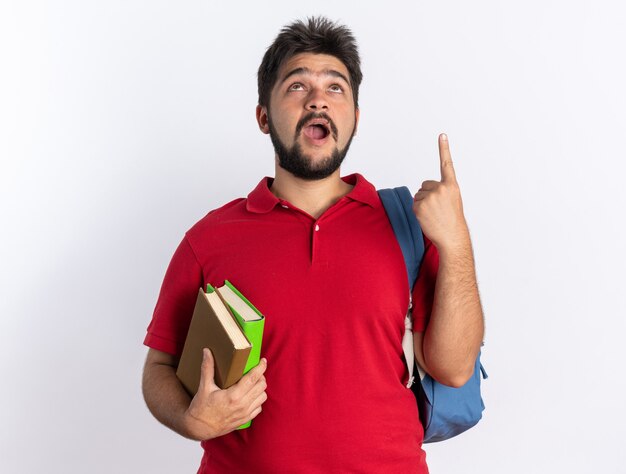 Młody brodaty facet student w czerwonej koszulce polo z plecakiem trzymającym notebooki patrzący w górę szczęśliwy i podekscytowany, wskazując palcem wskazującym w górę, stojąc nad białą ścianą