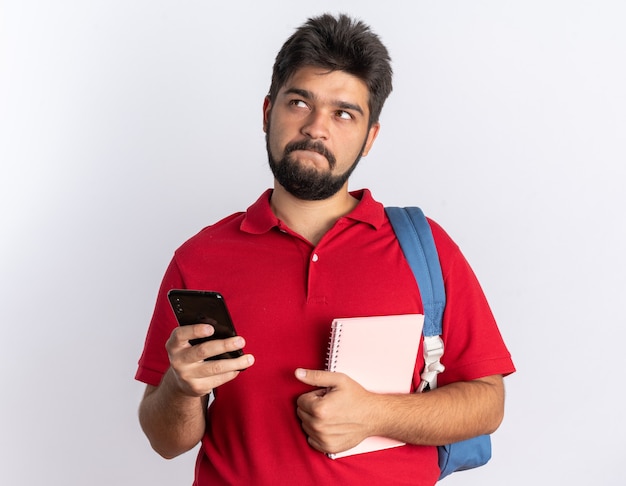 Młody, brodaty facet student w czerwonej koszulce polo z plecakiem trzymającym smartfona i notebooka, patrząc zdziwiony, stojąc nad białą ścianą