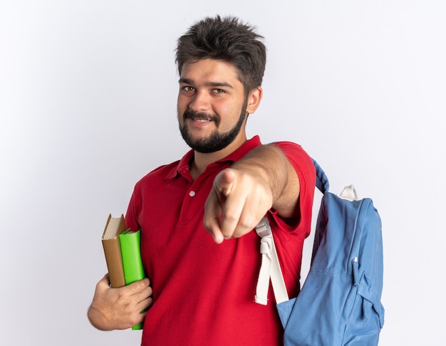 Młody brodaty facet student w czerwonej koszulce polo z plecakiem trzymającym zeszyty wskazującym palcem wskazującym uśmiechający się radośnie szczęśliwy i pozytywny stojąc nad białą ścianą
