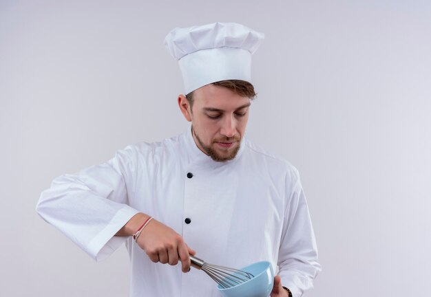 Młody brodaty mężczyzna kucharz ma na sobie biały mundur kuchenki i kapelusz trzyma łyżkę miksera na niebieskiej misce na białej ścianie