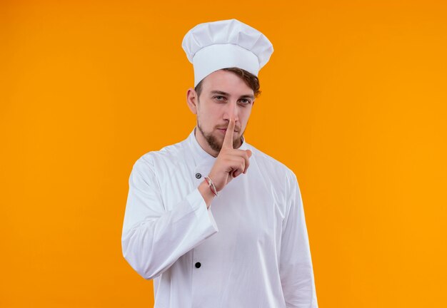 Młody brodaty mężczyzna kucharz w białym mundurze pokazując gest shh z palcem wskazującym na pomarańczowej ścianie