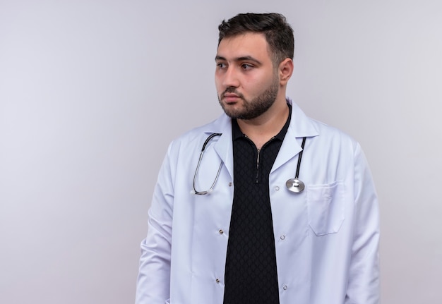 Młody brodaty mężczyzna lekarz ubrany w biały fartuch ze stetoskopem patrząc na bok z poważnym wyrazem pewności