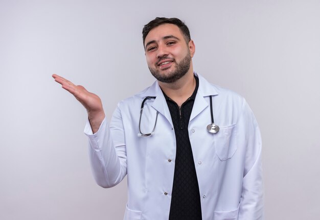 Młody brodaty mężczyzna lekarz ubrany w biały fartuch ze stetoskopem, przedstawiając ramieniem jego dłoni z uśmiechem