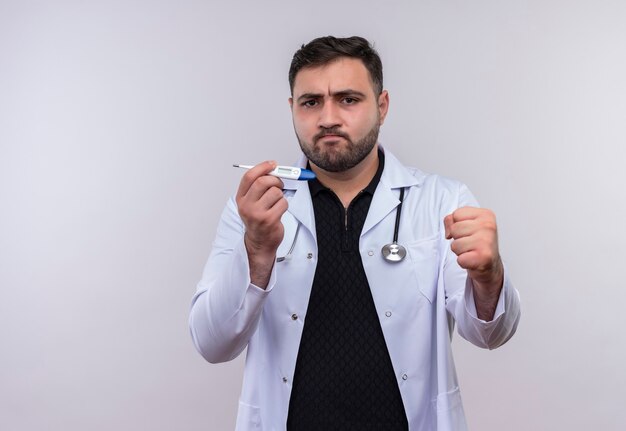 Młody brodaty mężczyzna lekarz ubrany w biały fartuch ze stetoskopem trzymając cyfrowy termometr uśmiechnięty pewny siebie zaciskając pięść, koncepcja zwycięzcy