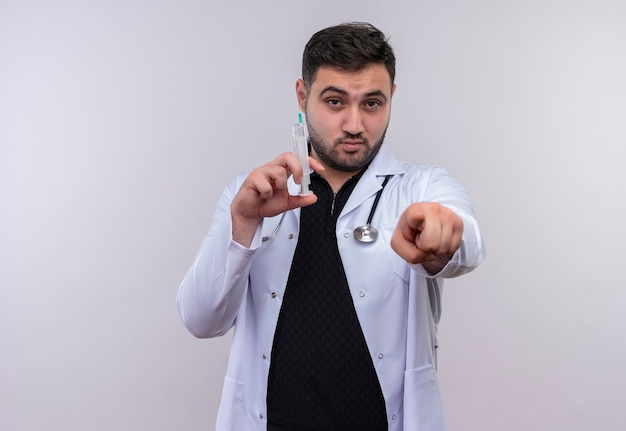 Bezpłatne zdjęcie młody brodaty mężczyzna lekarz ubrany w biały fartuch ze stetoskopem trzymając strzykawkę wskazując palcem na aparat z poważną twarzą