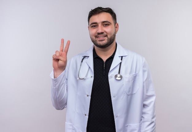 Młody brodaty mężczyzna lekarz ubrany w biały fartuch ze stetoskopem uśmiechnięty pokazując numer dwa lub znak zwycięstwa