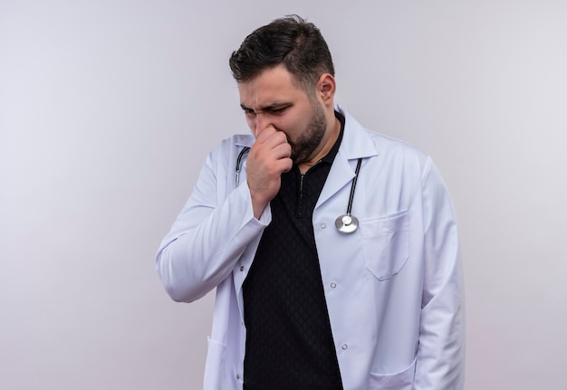 Młody brodaty mężczyzna lekarz ubrany w biały fartuch ze stetoskopem, zamykając nos palcami, czując nieprzyjemny zapach