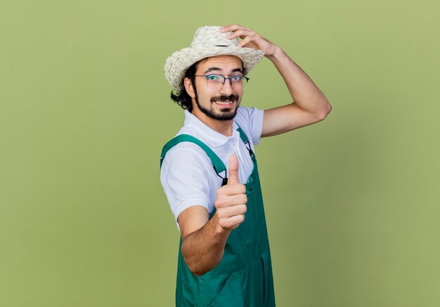 Młody brodaty mężczyzna ogrodnik na sobie kombinezon i kapelusz patrząc z przodu uśmiechnięty pokazując kciuki do góry stojąc nad jasnozieloną ścianą