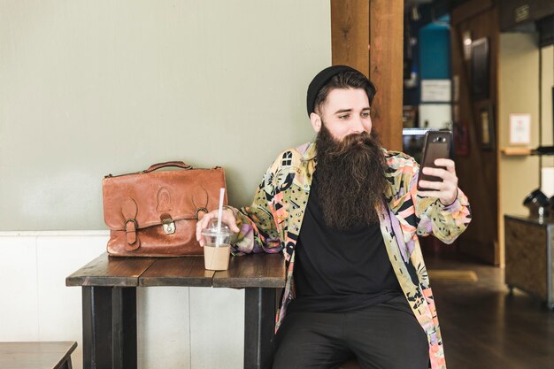 Młody brodaty mężczyzna siedzi w kawiarni przy selfie na telefon komórkowy