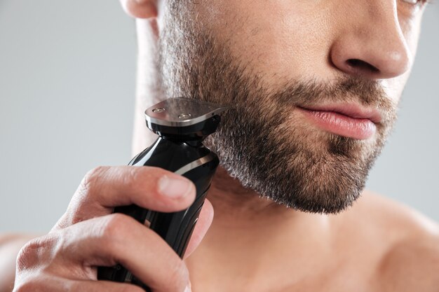 Młody brodaty mężczyzna używa elektryczną golarkę