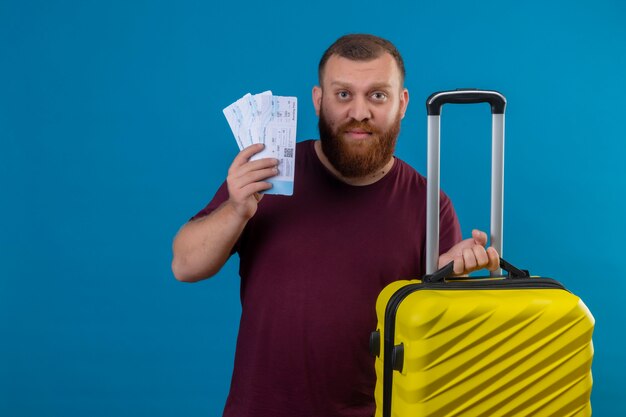 Młody brodaty mężczyzna w brązowej koszulce, trzymając walizkę podróżną i bilety lotnicze, patrząc na kamery ze smutnym wyrazem twarzy