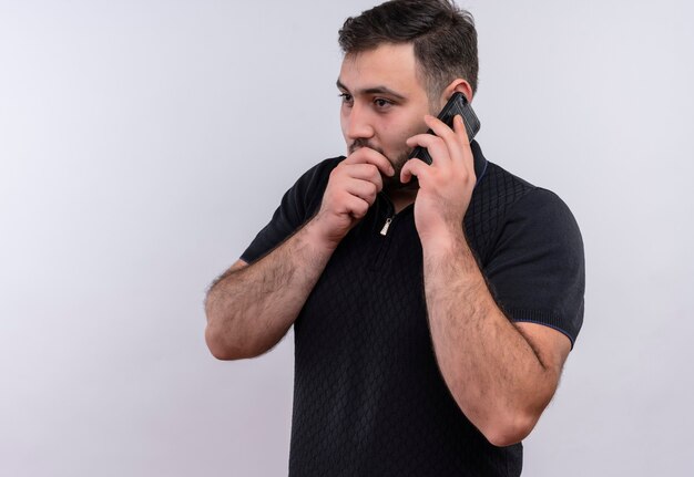 Młody brodaty mężczyzna w czarnej koszuli mówi sekret podczas rozmowy przez telefon komórkowy