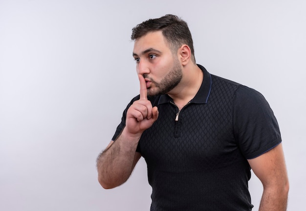 Młody brodaty mężczyzna w czarnej koszuli robi ciszy gest z palcem na ustach