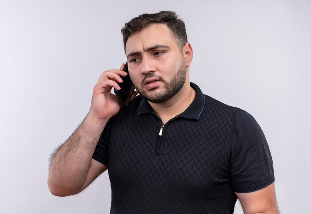 Młody brodaty mężczyzna w czarnej koszuli rozmawia przez telefon komórkowy z poważną twarzą