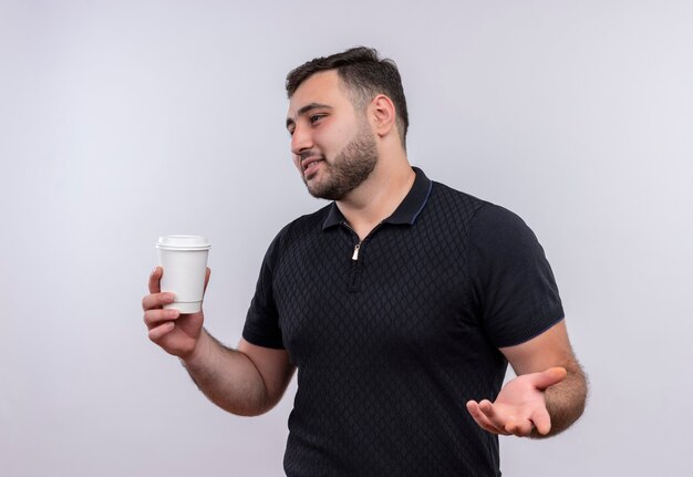 Młody brodaty mężczyzna w czarnej koszuli trzyma filiżankę kawy patrząc na bok gestykuluje ręką