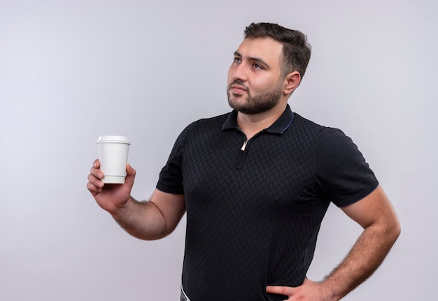 Młody brodaty mężczyzna w czarnej koszuli trzymając filiżankę kawy patrząc na bok z zamyślonym wyrazem twarzy