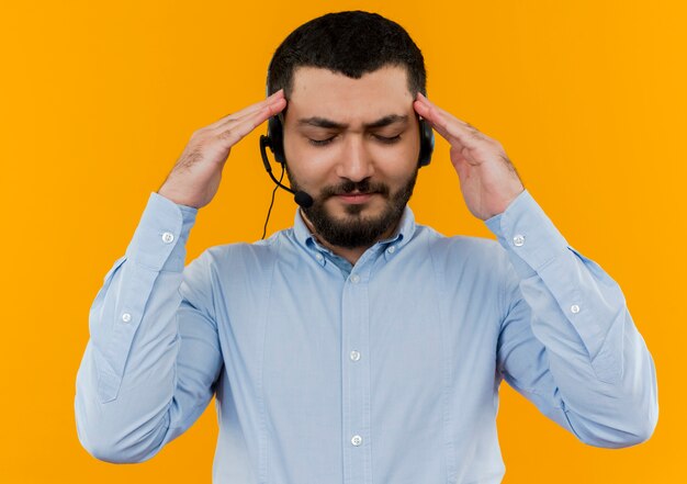 Młody brodaty mężczyzna w niebieskiej koszuli ze słuchawkami z mikrofonem, dotykając jego skroni o silnym bólu głowy