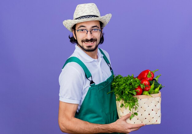 Młody brodaty ogrodnik mężczyzna ubrany w kombinezon i kapelusz trzymający skrzynię pełną warzyw patrząc z przodu uśmiechnięty wesoło stojąc nad niebieską ścianą
