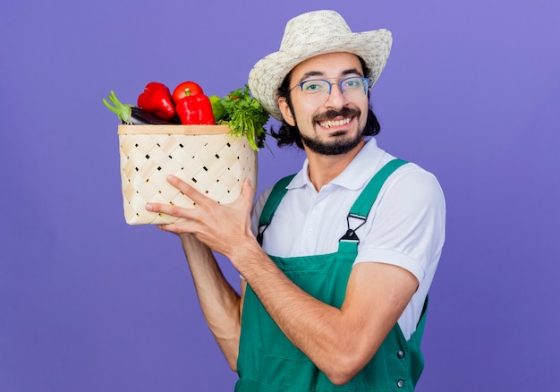 Młody brodaty ogrodnik mężczyzna ubrany w kombinezon i kapelusz trzymający skrzynię pełną warzyw patrząc z przodu uśmiechnięty ze szczęśliwą twarzą stojącą nad niebieską ścianą