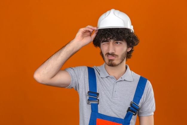 Bezpłatne zdjęcie młody brodaty przystojny budowniczy w mundurze konstrukcyjnym i kasku ochronnym, patrząc smutno i rozczarowany dotykając hełmu na odizolowanej pomarańczowej ścianie