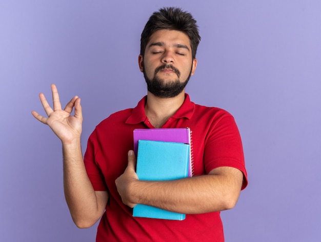 Młody Brodaty Student W Czerwonej Koszulce Polo, Trzymający Książki, Wykonujący Gest Medytacji Relaksujący Z Zamkniętymi Oczami, Stojący Nad Niebieską ścianą