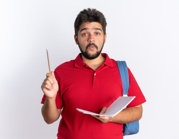 Młody, brodaty student w czerwonej koszulce polo z plecakiem trzymającym notatnik i ołówek, wyglądający na zdziwionego stojącego