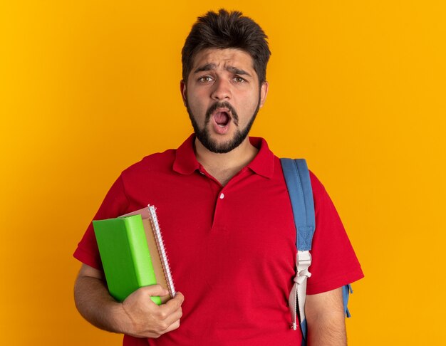 Młody, brodaty student w czerwonej koszulce polo z plecakiem trzymającym zeszyty, wyglądający na zdezorientowanego i zdziwionego?