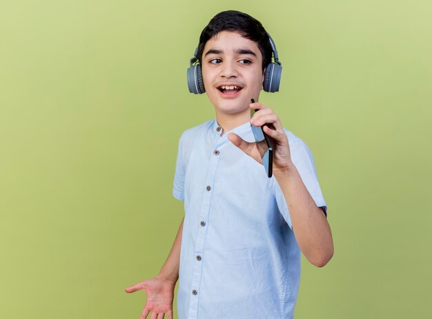 Młody chłopak noszenie słuchawek śpiewa patrząc na bok przy użyciu telefonu komórkowego jako mikrofonu na białym tle na oliwkowej ścianie