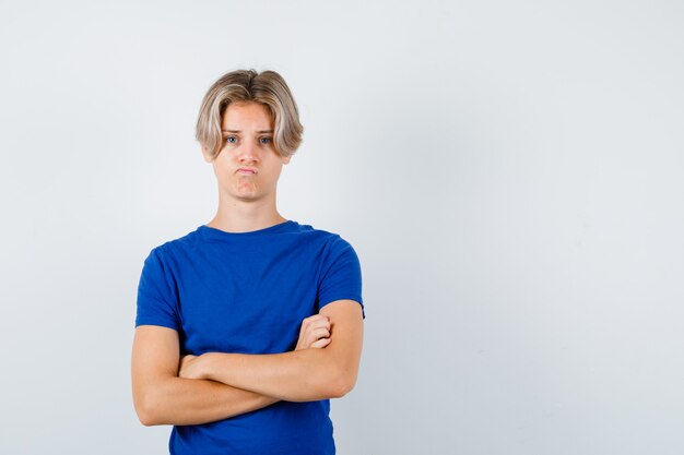 Młody chłopak teen w niebieskim t-shirt, trzymając założone ręce i patrząc niezadowolony, widok z przodu.