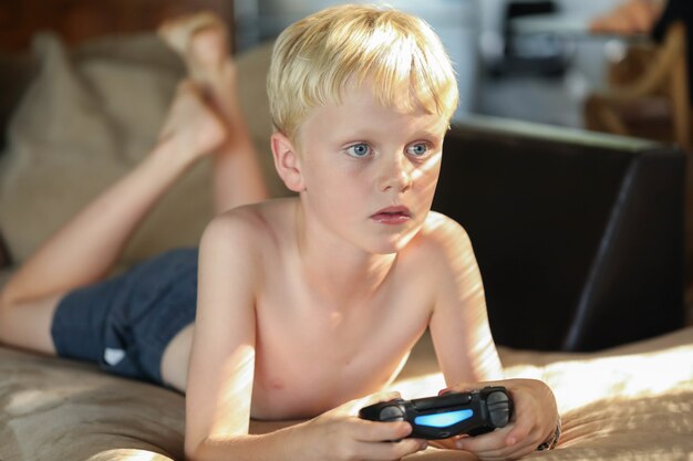 Młody chłopiec rasy kaukaskiej grający w gry wideo w domu na kanapie w miękkim popołudniowym świetle
