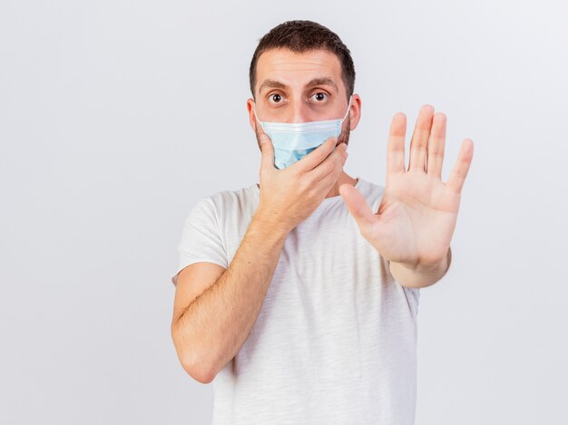 Młody chory człowiek ubrany w szalik i maskę medyczną zawinięty w kratę pokazujący gest stop na białym tle