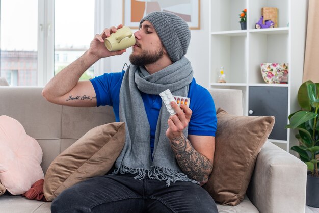 młody chory mężczyzna ubrany w szalik i czapkę zimową siedzący na kanapie w salonie, trzymający paczki leków medycznych popijający filiżankę herbaty